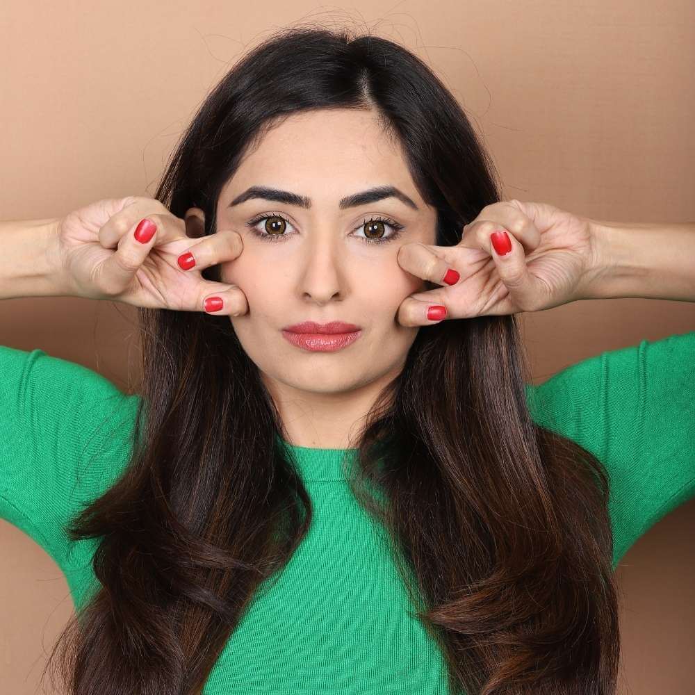 A young woman in a green t-shirt displaying a face yoga technique with 2 fingers placed on each side of her cheeks for a younger and toned looking skin. 