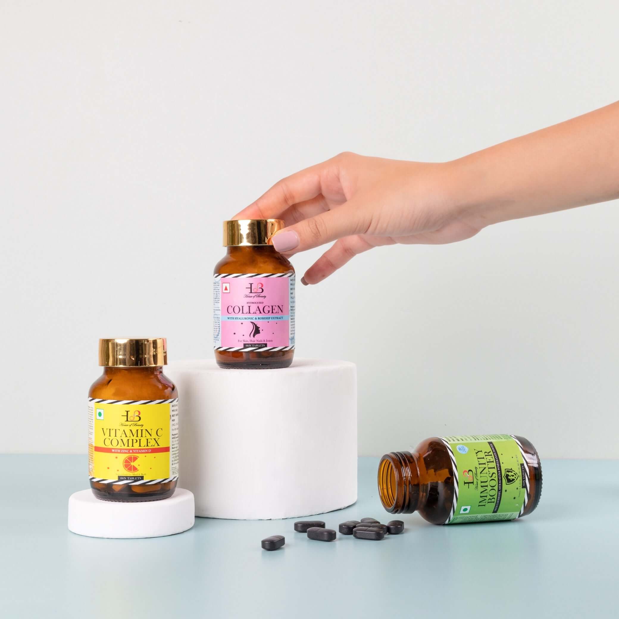 Three bottles of hair and skin supplements on a white background, with two bottles on white cylindrical blocks and one laying flat. The bottles have yellow, pink, and green labels reading VITAMIN C COMPLEX, COLLAGEN, and IMMUNITY BOOSTER.