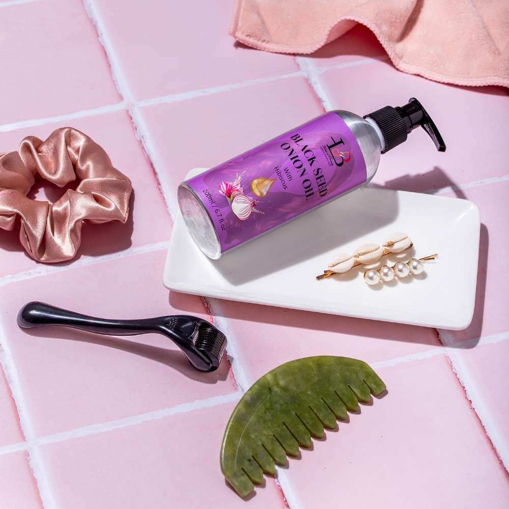 Flat lay of hair care items on a pink tiled surface, including a bottle of Black Seed Onion Oil for hair fall reduction, a jade comb for relaxation, a hair scrunchie, a derma roller for collagen boost and wrinkles, at House of Beauty India.