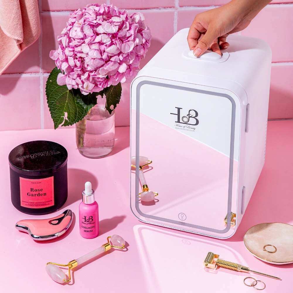 A hand-holding mini white skincare fridge with the logo House of Beauty with skincare products- Rose Quartz roller, Collagen serum bottle, and Gua Sha for fine lines, anti-aging, and collagen boost with some props like flowers and rings lying flat.