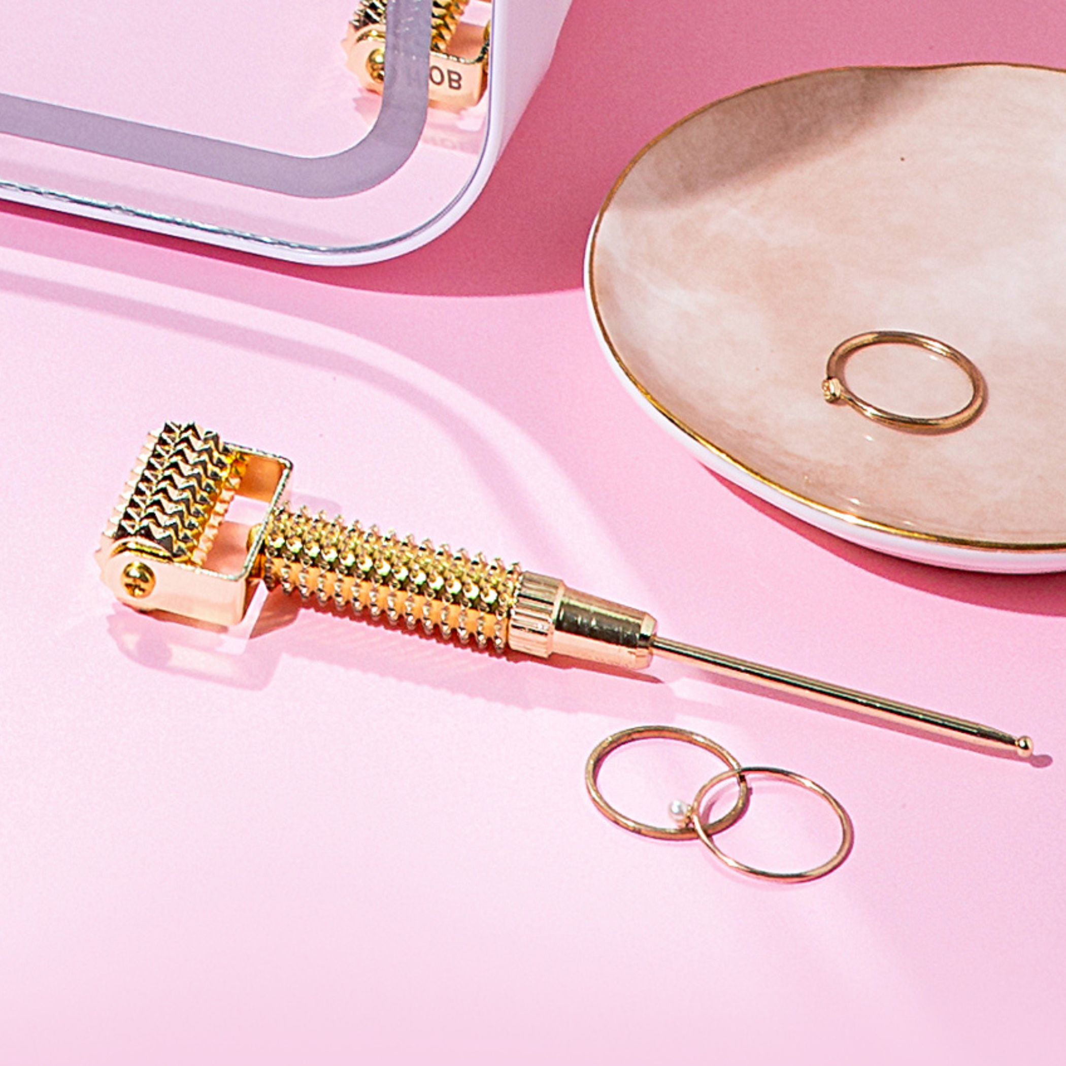 A gold collagen roller with lying flat on a pink surface by House of Beauty used for wrinkles, collagen boost and anti-aging.